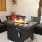 A black wicker fire table with a glass top and visible gas valve, set against a white background.