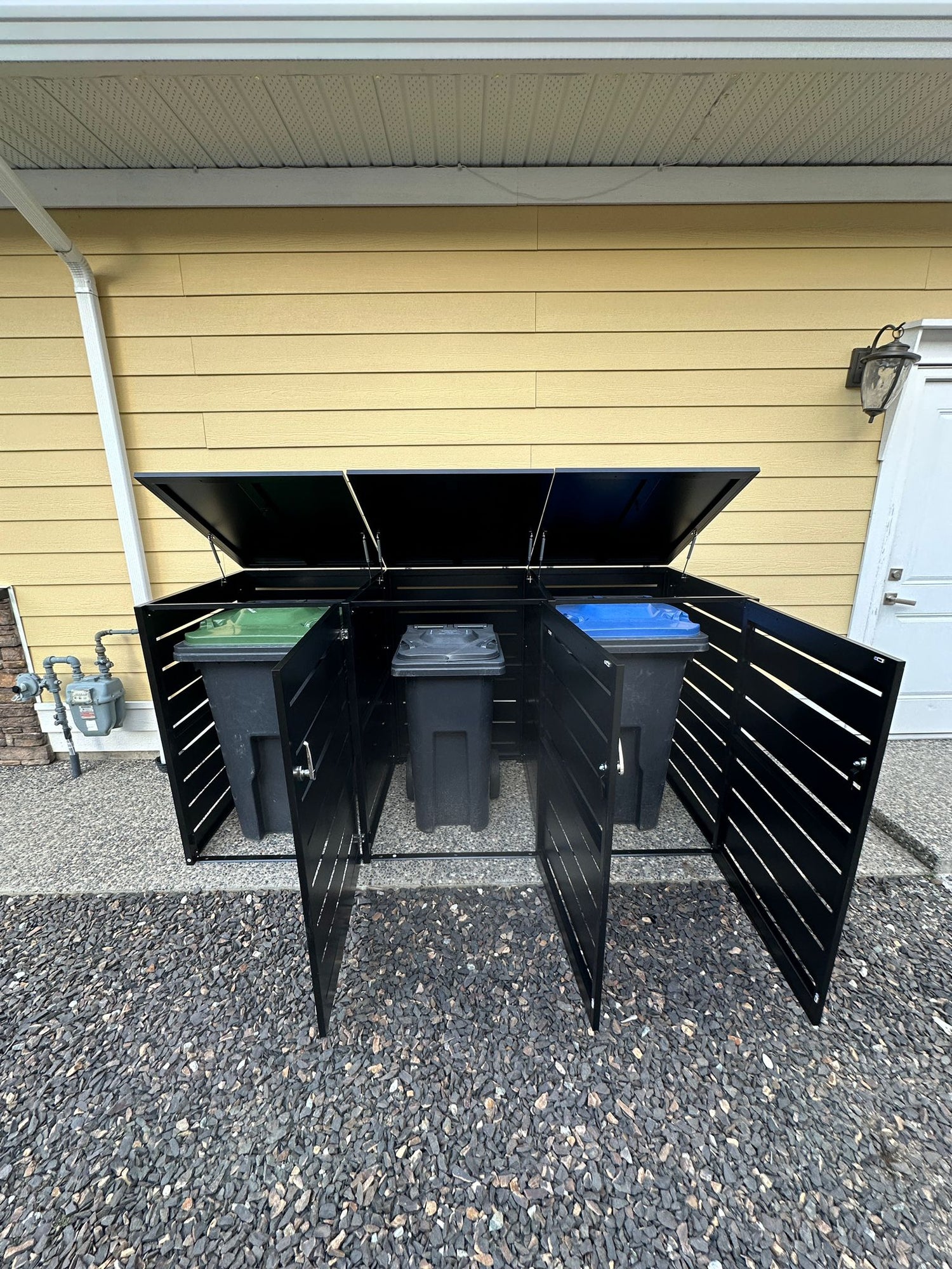 Black outdoor storage unit with open doors revealing trash bins against a yellow wall.